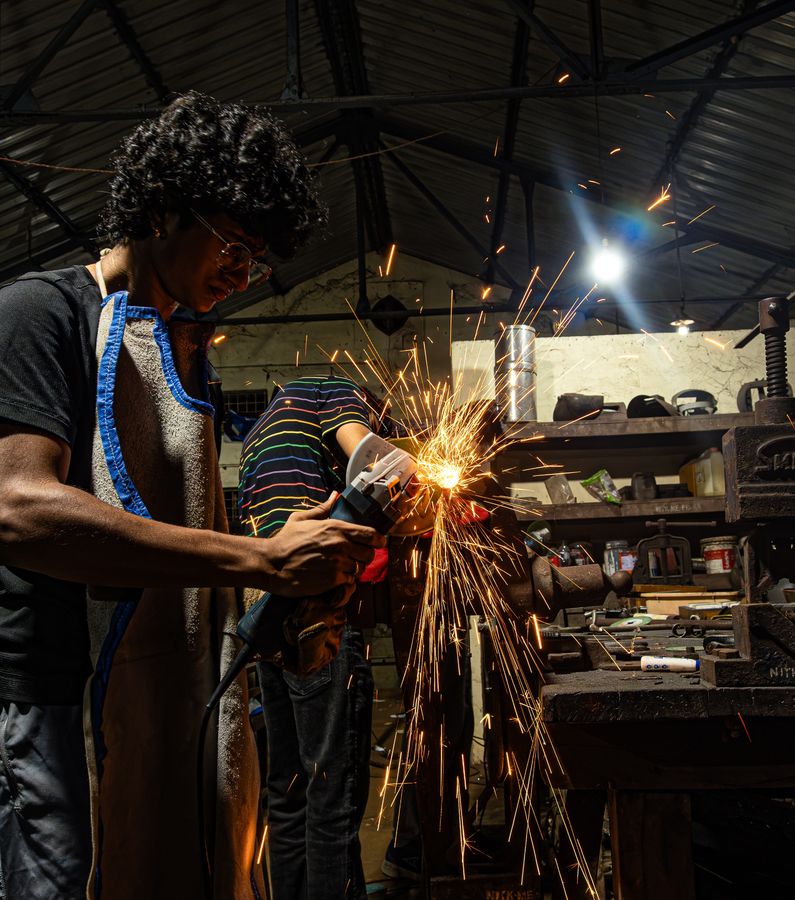 A team member in a leather apron cutting metal with an angle grinder, sparks spraying across the workshop